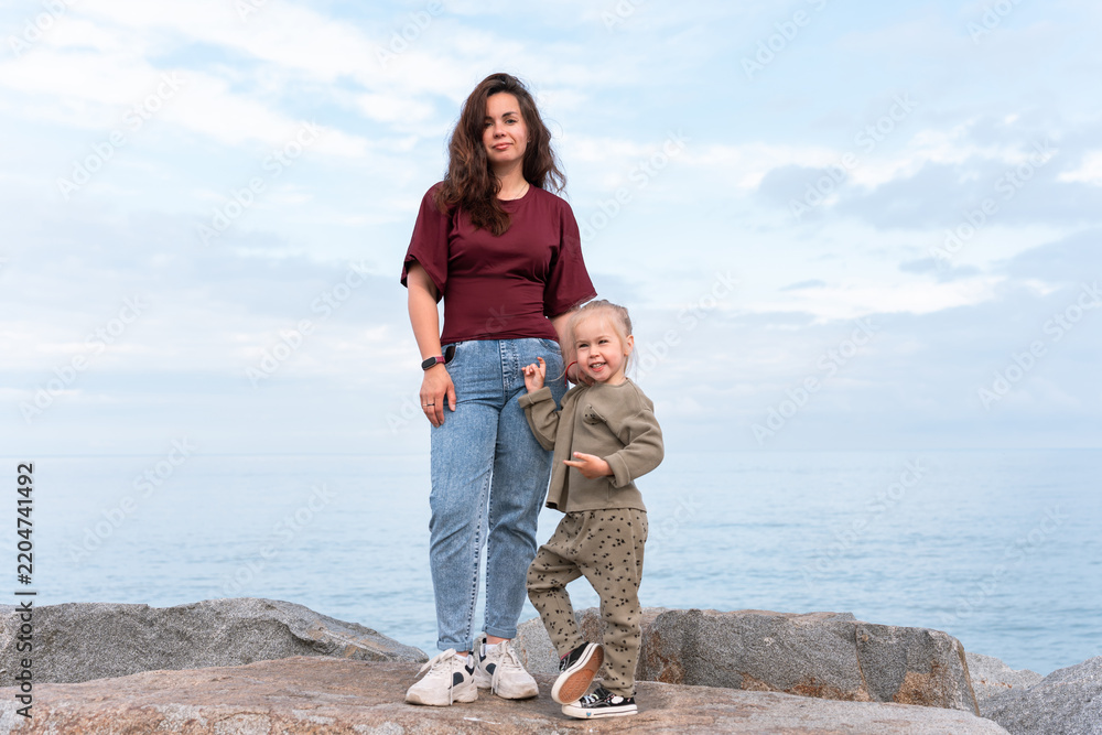 Obraz premium A young mother standing by the sea with her little daughter. The woman smiles gently as the child hugs her, with calm water and a cloudy sky in the background. Family, love, and lifestyle concept.
