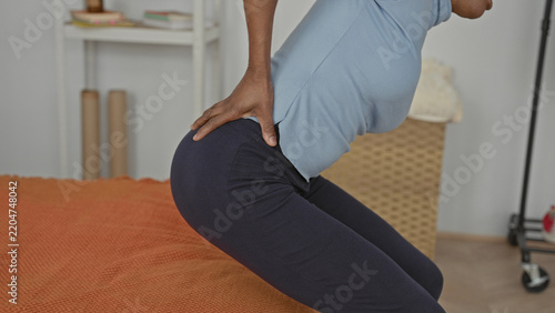 Woman sitting on bed holding lower back and leaning forward on orange bedcover in building, visible hip and hand contact with back; pain recovery.