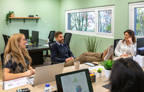 Obraz na plátně Seated man chats with smiling women while resting feet on their shared desk
