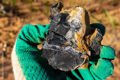 Raw black flint shard with sparkling quartz veins held by a hand in a green work glove, showing geological mineral texture in a natural outdoor environment