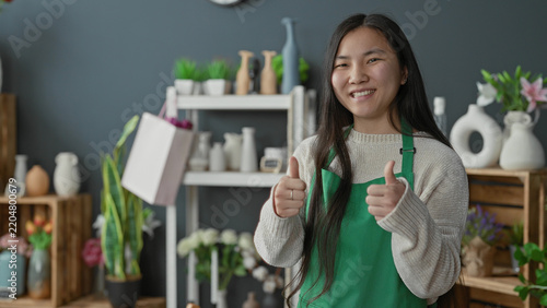 фотографія Woman florist in green apron gives thumbs up with both hands in studio surrounded by flowers and vases on shelves; local success happiness