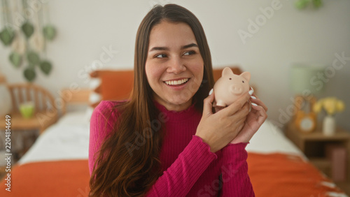 Konstfotografi Woman holds piggy bank to cheek on bed in bedroom, smiling and looking aside while wearing pink sweater near orange blanket and bedside lamp; saving hope
