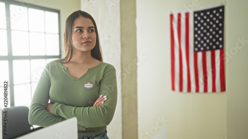 Fotografie Woman with arms crossed wearing a voted sticker at a polling station beside an american flag in a building; civic pride