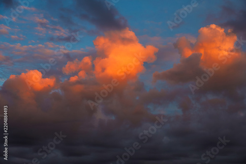 Beautiful sunset sky filled with dramatic cumulus clouds illuminated in vibrant orange and golden colors contrast against a deep blue and dark grey background
