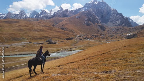 Aerial rear follow shot of traveler horseback riding across golden mountain valley towards snow-capped peaks near Kel-Suu Lake, Kyrgyzstan