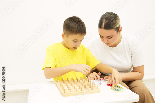 Boy stretches colored rubber bands around pegs to reproduce a geometric pattern from a reference card while a specialist guides the educational activity.