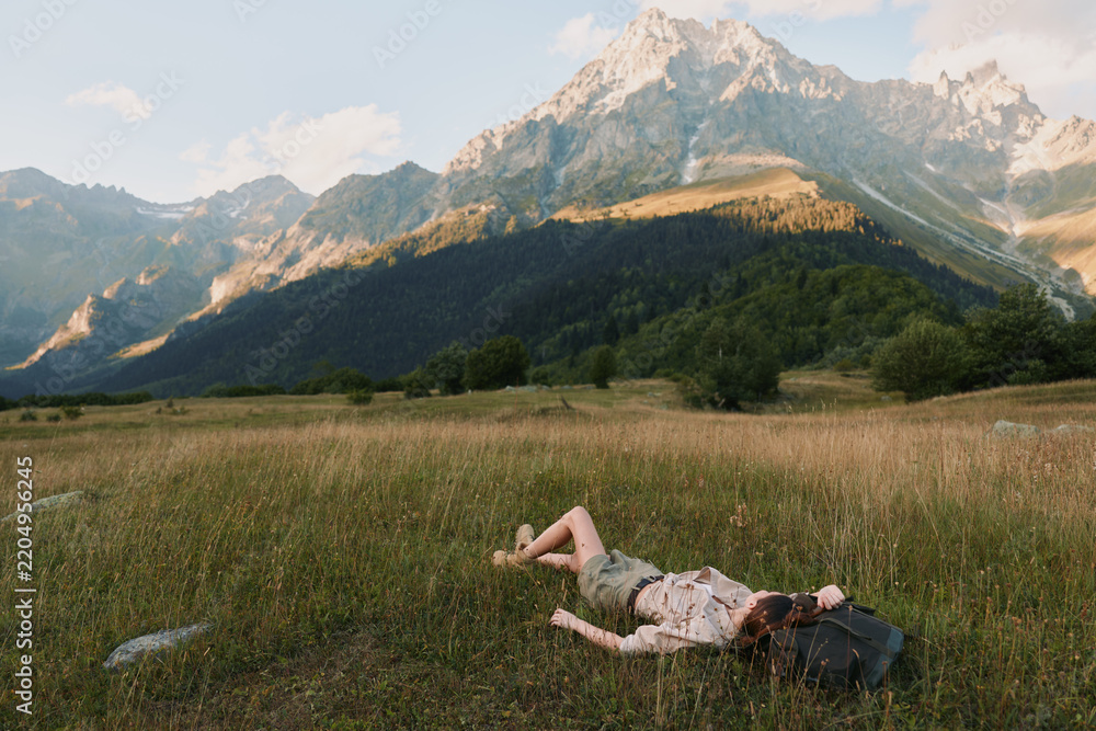 Obraz premium Young woman lies on a grassy field in casual clothes resting with a backpack near her head against a backdrop of sunlit mountains under a partly cloudy sky.