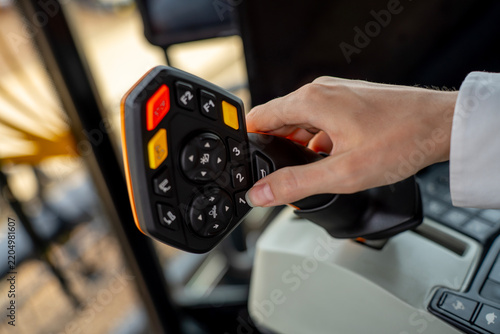 Operator hand holding multifunctional control joystick with buttons inside modern agricultural tractor or combine harvester cabin.