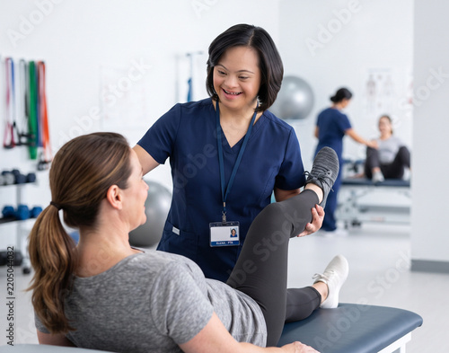 Friendly female physical therapist assistant with Down syndrome assisting a patient with leg stretches in a clinic