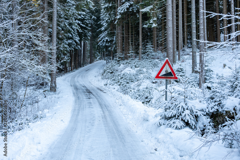 Obraz premium Road sign warning of a steep incline beside an icy and snow covered road in a winter landscape, highlighting hazardous driving conditions.