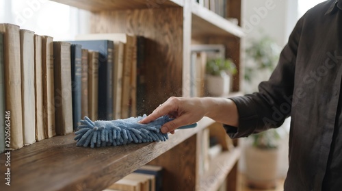 A person's hand meticulously dusting a wooden bookshelf filled with old books and decorative items, highlighting home cleaning and maintenance.