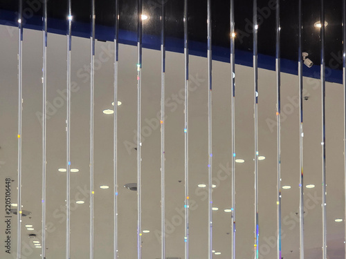 A pattern of vertical glass rods forming a decorative partition with colorful light refractions. Reflections of bright ceiling spotlights on a glossy surface create a modern architectural background.