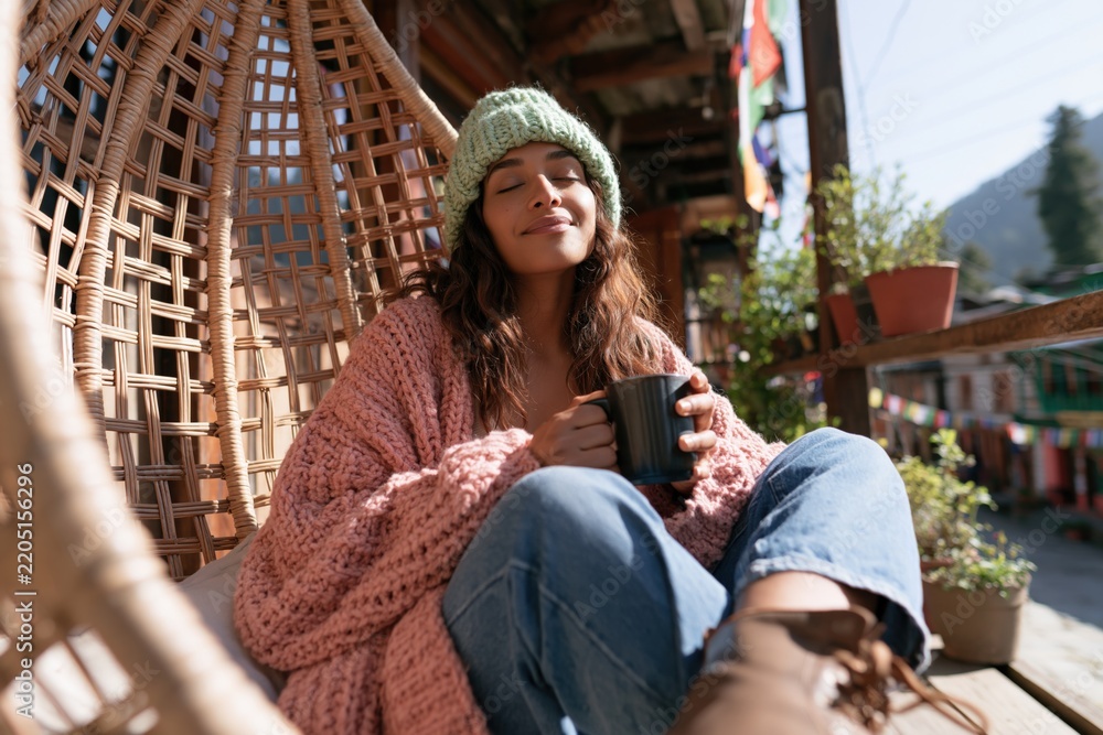 Obraz premium Smiling woman drinking coffee in a wicker swing chair on a sunny balcony for cozy lifestyle wallpaper and travel relaxation background