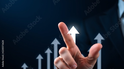 A businessman in a suit pointing at a series of glowing white arrows that are trending upwards on a dark blue background, for business growth