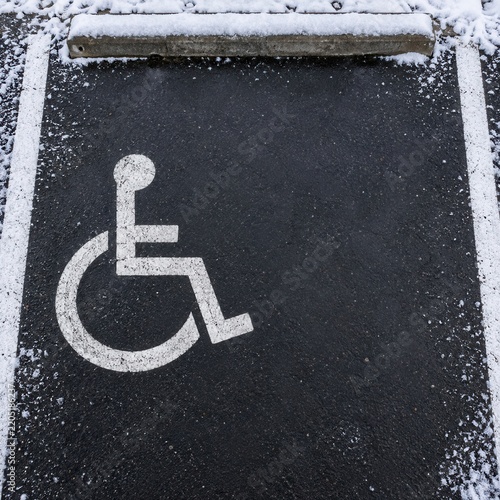 Accessible parking space with white wheelchair symbol painted on dark asphalt dusted with snow, top view