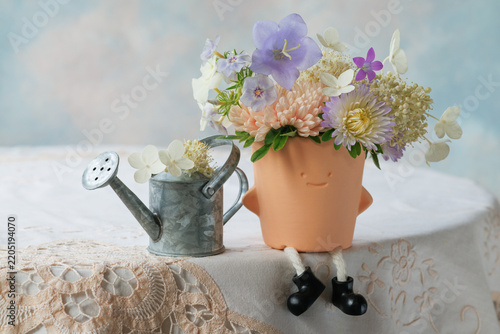 Whimsical still life on a table with a lace tablecloth: a watering can with flowers and a decorative flower pot featuring a smiling face and boots; asters, hydrangea, bellflowers, and phlox.