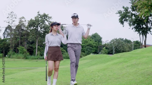 Young Asian couple carrying golf clubs walk together across green course during coaching session