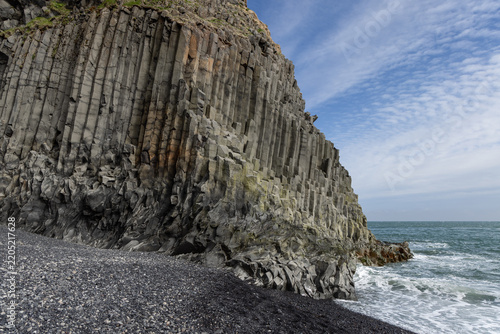 Towering basalt columns rise above black pebbles and Atlantic surf at Reynisfjara Beach in Iceland, revealing rugged volcanic geometry beneath a blue, clouded sky.