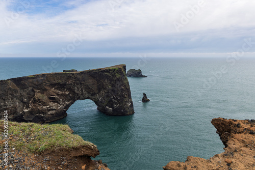 Wide coastal view of Dyrholaey Arch, a massive natural opening in rugged volcanic sea cliffs, with offshore rock formations and the Atlantic Ocean in South Iceland.
