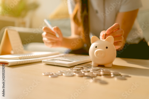 Woman putting a coin into a piggy bank while using a calculator and notebook, representing saving money, budgeting, personal finance and financial planning at home