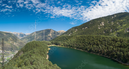Aerial view of Lake Engolasters surrounded by dense pine forests and mountains with radio towers under a blue sky in Encamp, Andorra.