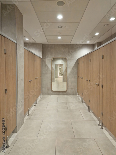 Modern public restroom corridor with wooden cubicle doors, gray tiled floor, and a full-length mirror