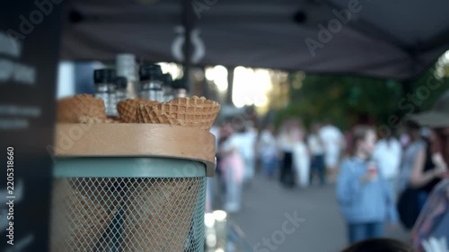 Close up shows crispy empty waffle cones resting in metal mesh holder at ice cream stall. Defocused passersby enjoy walking through sunny public park in blurred background.