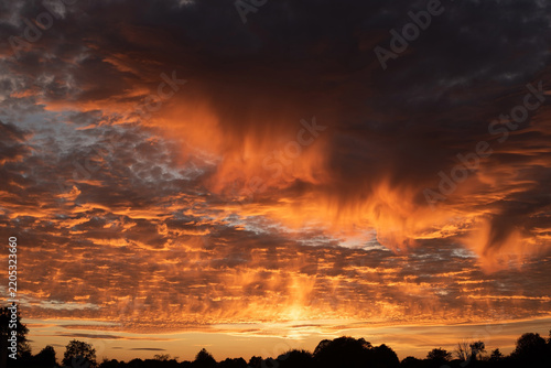A dramatic orange sunset lights textured clouds and falling rain shafts above a silhouetted treeline, creating fiery, moody landscape. The photo is a little out of focus but really beautiful