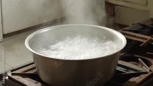 Closeup static shot of metal pot vigorously boiling with rising steam on gas burner over stove in home kitchen interior countertop