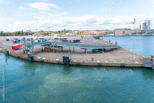 Canvastavla Elevated loading ramp dominates a large ferry terminal beside harbor water, with freight trailers, docking equipment and city buildings beyond