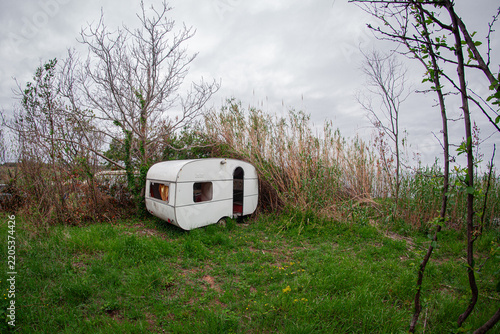 Картината върху платно Abandoned vintage caravan trailer with broken windows, missing door panel, and rust damage, sitting in overgrown grass among trees, showing signs of vandalism and decay