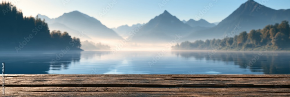 Obraz premium Wooden table surface lies in front of a blurred mountain lake at sunrise. Mist rises off the water reflecting nearby mountains.