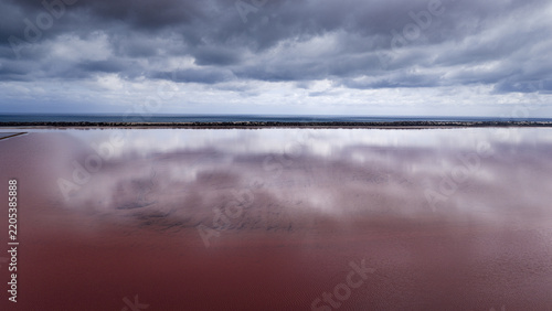 Aerial view of a vibrant pink salt lake with dramatic dark clouds reflecting on the water surface and a distant coastline in Australia.