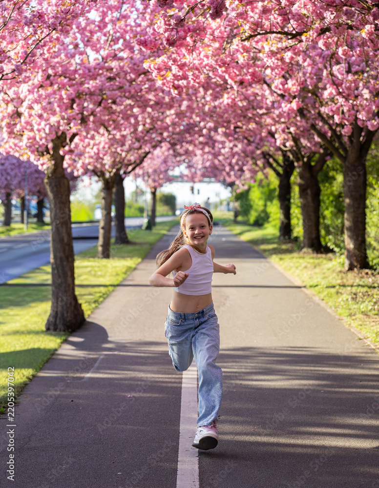 Obraz premium Happy girl running under blooming cherry blossom trees on sunny spring avenue