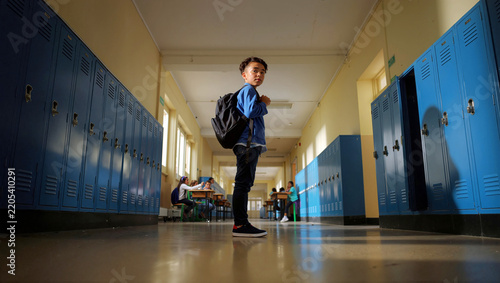 A focused male student wearing a backpack stands in a school hallway lined with blue lockers, looking back with a ready and determined expression before his next class starts.