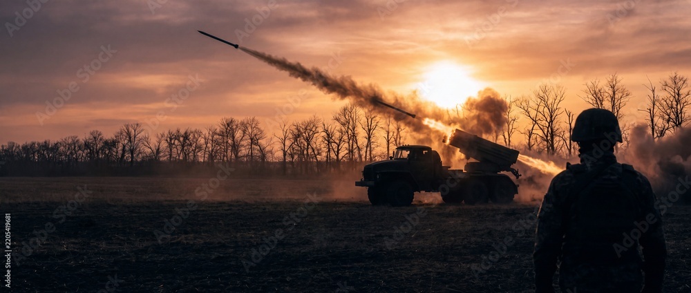 Obraz premium Soldier observing a multiple rocket launcher system releasing rockets in a fiery explosion against a dramatic sunset sky