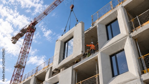 Construction workers lift a large concrete panel into place at a building site with a crane on a sunny day