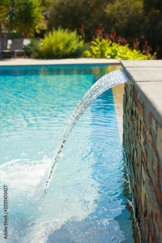 Elegant swimming pool with a cascading water feature flowing from a stone wall into clear turquoise water, surrounded by lush Mediterranean garden vegetation. Bright sunny outdoor scene with a relaxin