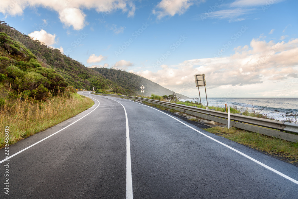 Fototapeta premium Empty winding coast road under blue sky with clouds at sunset in autumn. A thin mist is drifting from the ocean.