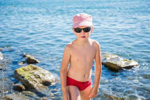 Young blond boy in pink striped cap and black sunglasses stands sadly in shallow blue sea near mossy rocksю