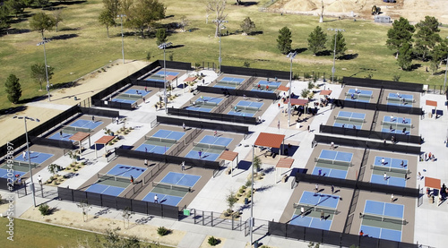 Aerial view of several rows of pickleball courts with players on the blue and green surfaces, surrounded by fencing and park grounds in Valencia, California, United States.