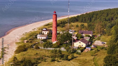 Red lighthouse on sandy baltic sea coastline with lush green forest and summer beach landscape.