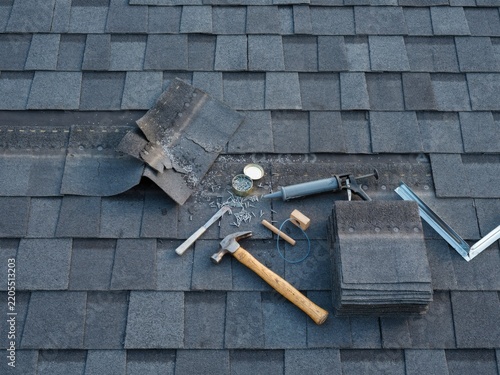Roofing tools and dark grey shingles on a damaged roof ready for repair and replacement.