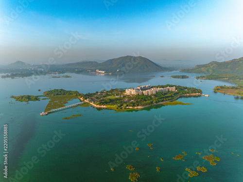 Aerial view of the Taj Lake Palace situated on an island in Lake Pichola, surrounded by calm turquoise waters and distant hills in Udaipur, Rajasthan, India.