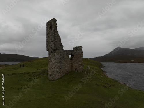 Aerial view of the ruins of a historic stone castle tower situated on a grassy headland beside a loch with distant mountains under a cloudy sky in Scotland, United Kingdom.