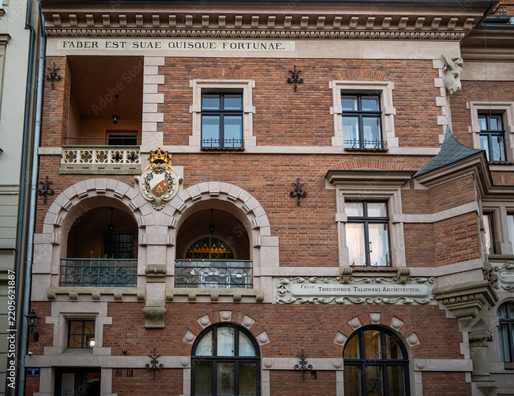 Obraz premium The detailed facade of a historical brick building showcases large arched openings, a balcony, and a prominent golden crest by architect Theodor Talowski in Krakow, Poland.