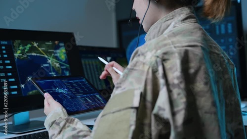 Asian female military officer in camouflage uniform analyzes radar maps on screens and writes notes in a high-tech tactical command center.