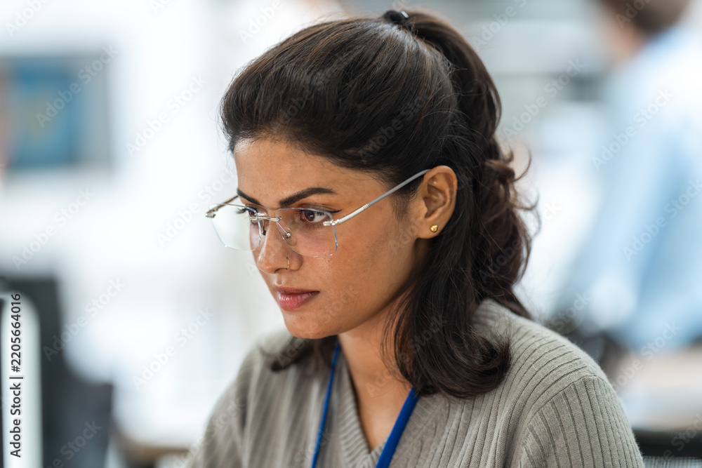 Obraz premium Close-up of Indian female engineer with glasses and ID lanyard focused on her work in a bright office, in an unposed working moment, an inclusive technology workplace scene