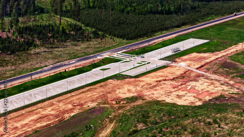 Aerial view of a paved parking area and rest stop situated alongside a road, bordered by dense forest and cleared earth in Latvia.