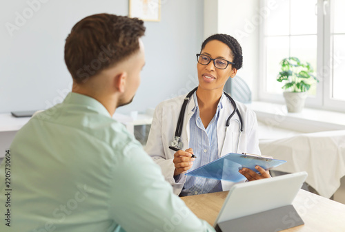 Portrait of woman physician listening and writing down patient's complaints sitting at the desk in clinic during medical examination. Sick man visiting a doctor. Healthcare and medicine concept